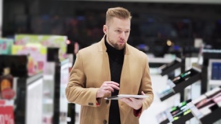 年轻男子穿着棕色外套，手持数字平板，在购物中心内行走，稳定器拍摄视频素材模板下载