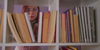 深思熟虑的戴眼镜的女人站在书架后面看着书。年轻的