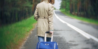 年轻美丽的女子站在路边，手持一个行李箱，想要搭便车