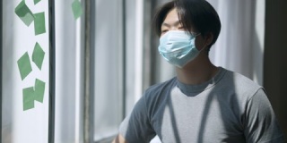 年轻英俊的亚洲男子戴着冠状病毒面罩站在窗口，脸上粘糊糊的