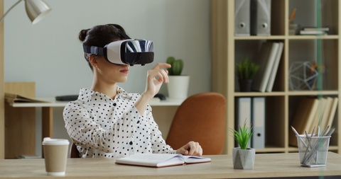 高加索人女性戴着VR眼镜坐在办公桌前，滚动或进行其他操作