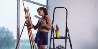 年轻的画家站在大窗户旁边的艺术工作室里画画。