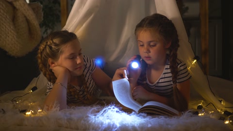 幸福的家庭。两个女儿在帐篷里看书。手里拿着手电筒视频素材模板下载