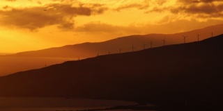 在群山和海洋的背景下，夕阳照亮了风力涡轮机