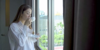 穿着睡衣站在窗边喝茶的女人的侧视图